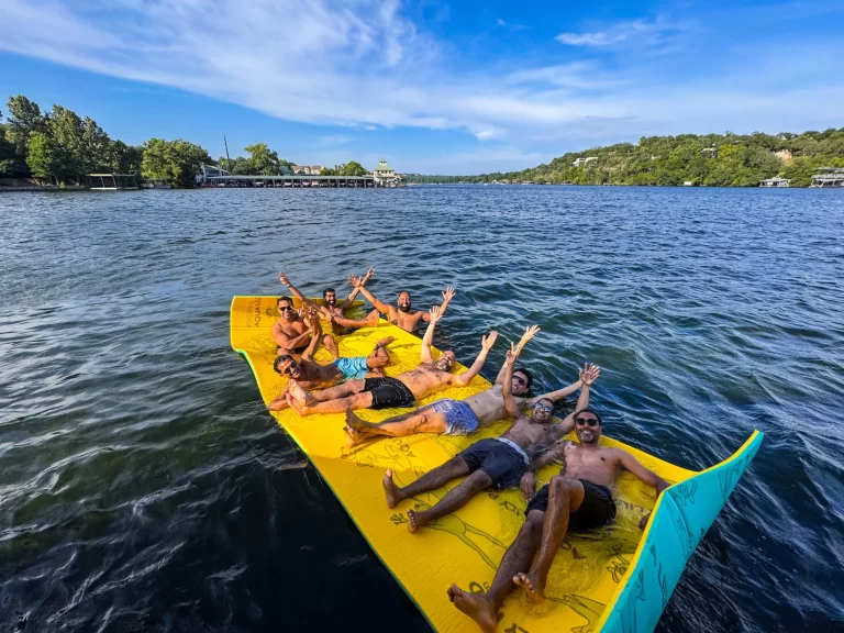 Group of friends relaxing and raising their arms while floating on a large pad during a Lake Austin boat rental outing